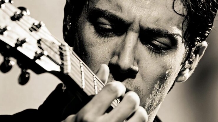 Uomo concentrato in primo piano che tiene il manico della chitarra, preme e pizzica le corde sul palco, con camicia scura e monitor in-ear.