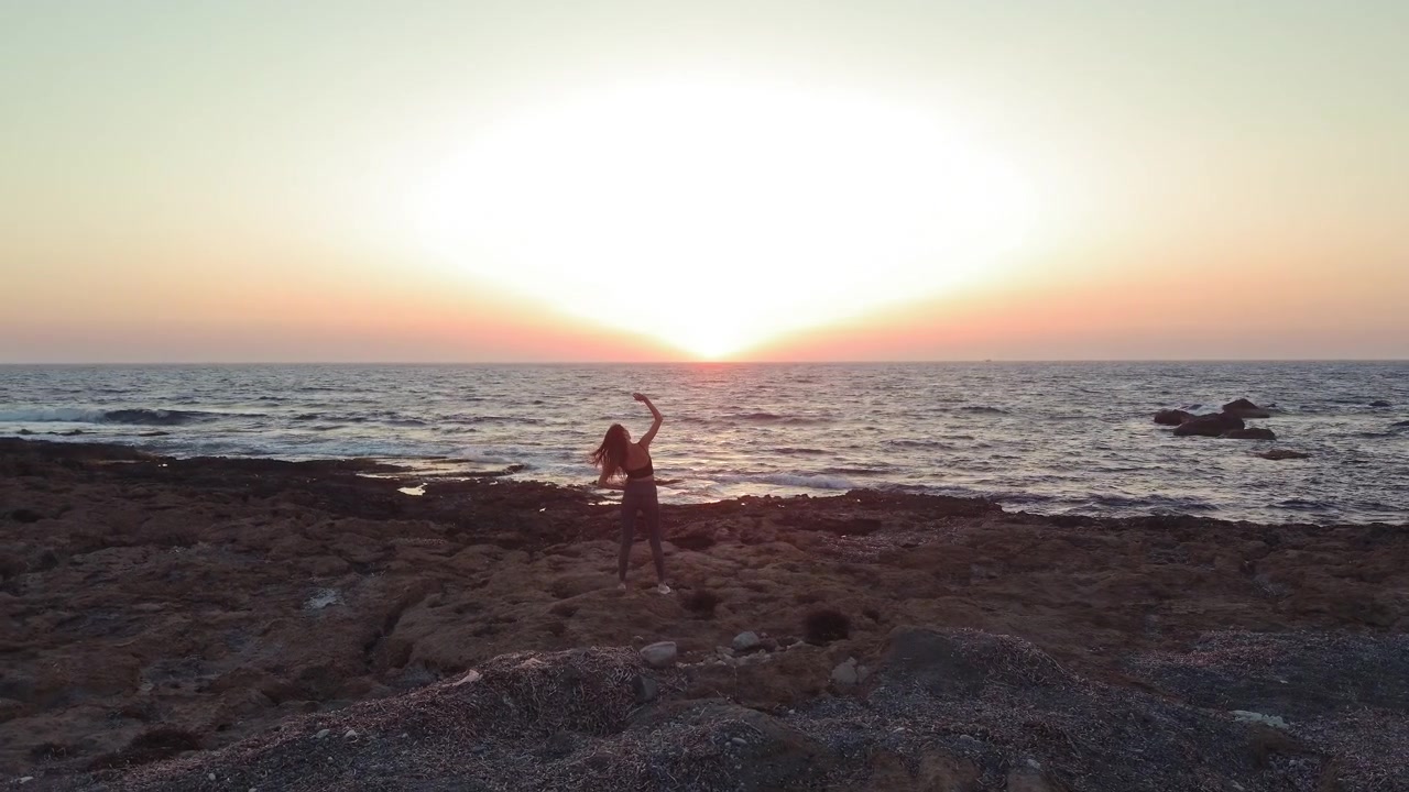 Una donna esegue un esercizio di stretching su una costa rocciosa e frastagliata mentre il sole tramonta sulla calma