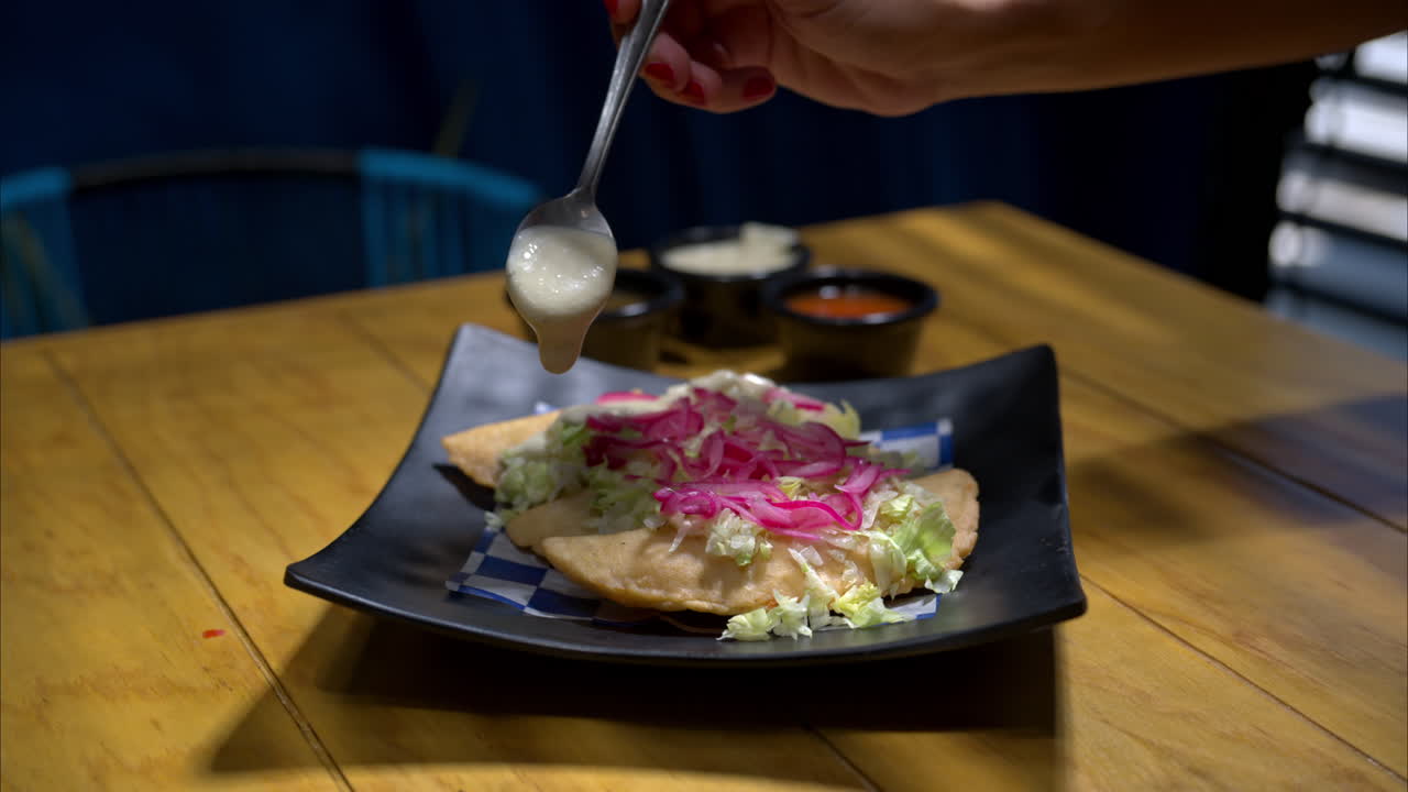 movimiento lento de una mujer vertiendo crema agria en un plato de empanadas tradicionales mexicanas servidas con lechuga y cebolla roja