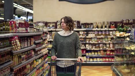 Portrait-of-a-woman-with-wavy-hair,-wearing-casual-is-driving-shopping-trolley-through-the-row-with-full-of-goods-shelf-in