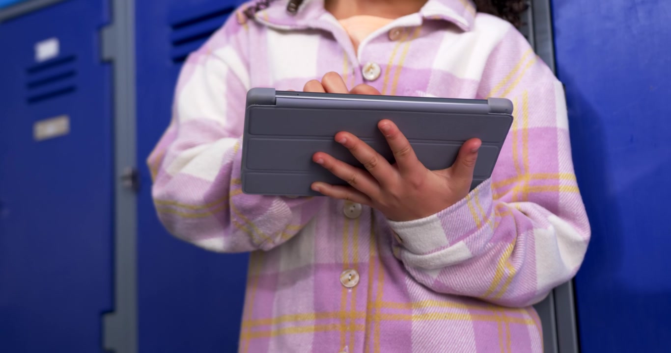 African American teenage girl leaning at lockers in plaid coat checking messages on gray tablet