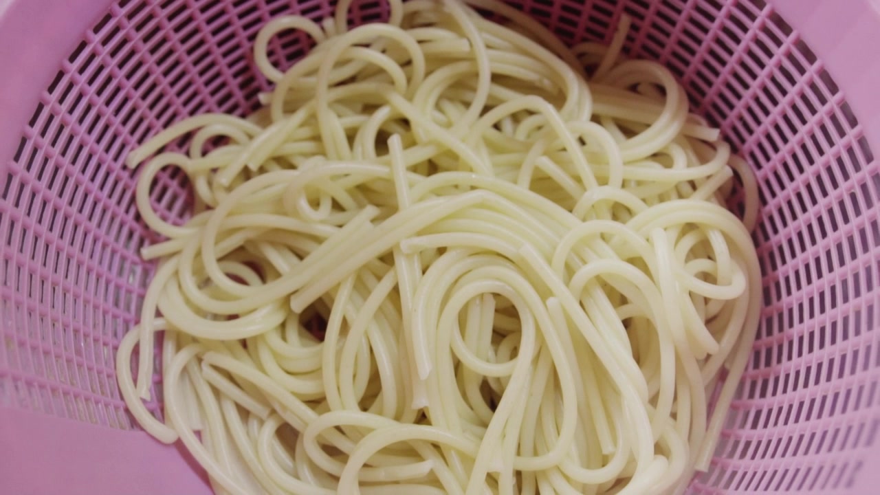 Primo piano di spaghetti cotti che scolano in uno scolapasta rosa con vapore visibile che sale. Adatto per tutorial di cucina, cucina italiana e video di preparazione dei pasti.