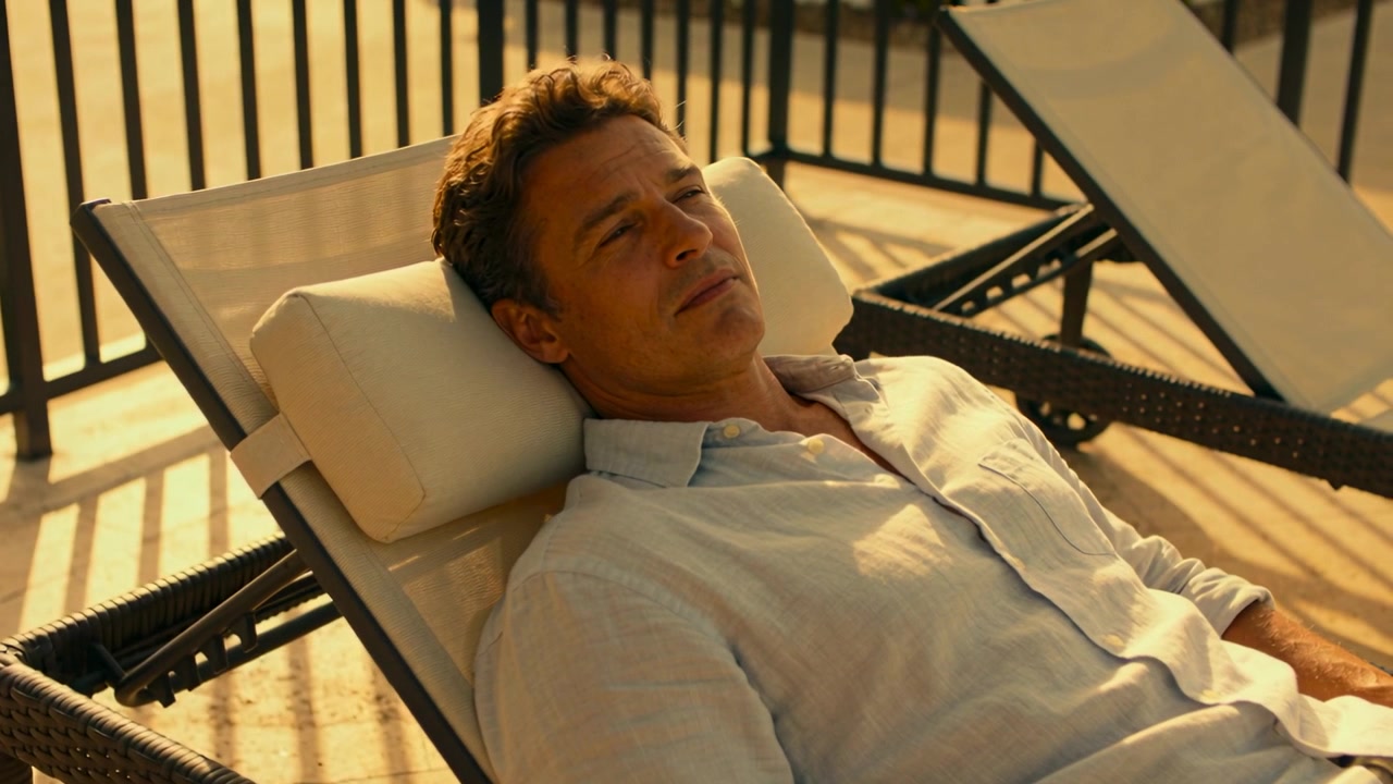 Shifting sunlight nudging man in linen shirt, lifting head and opening eyes on chaise with pillow