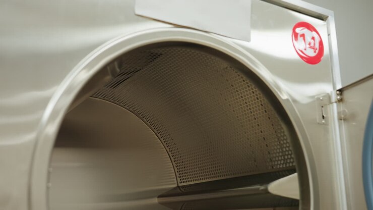 Inner view of industrial washer with door open showing stainless steel drum surface and control panel details inside modern laundry facility with bright lighting and reflections on metallic surface