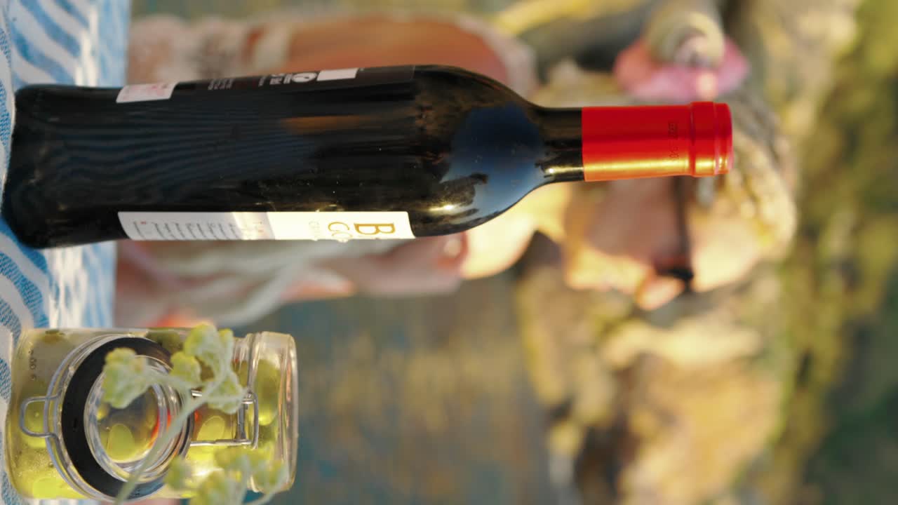 Ripresa verticale di un picnic in spiaggia con vino, olive e fiori gialli, sullo sfondo una giovane ragazza e il mare.