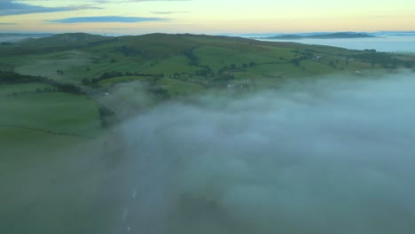 Flying-high-over-misty-M6-motorway-with-fog-bank-and-green-hills-at-sunrise