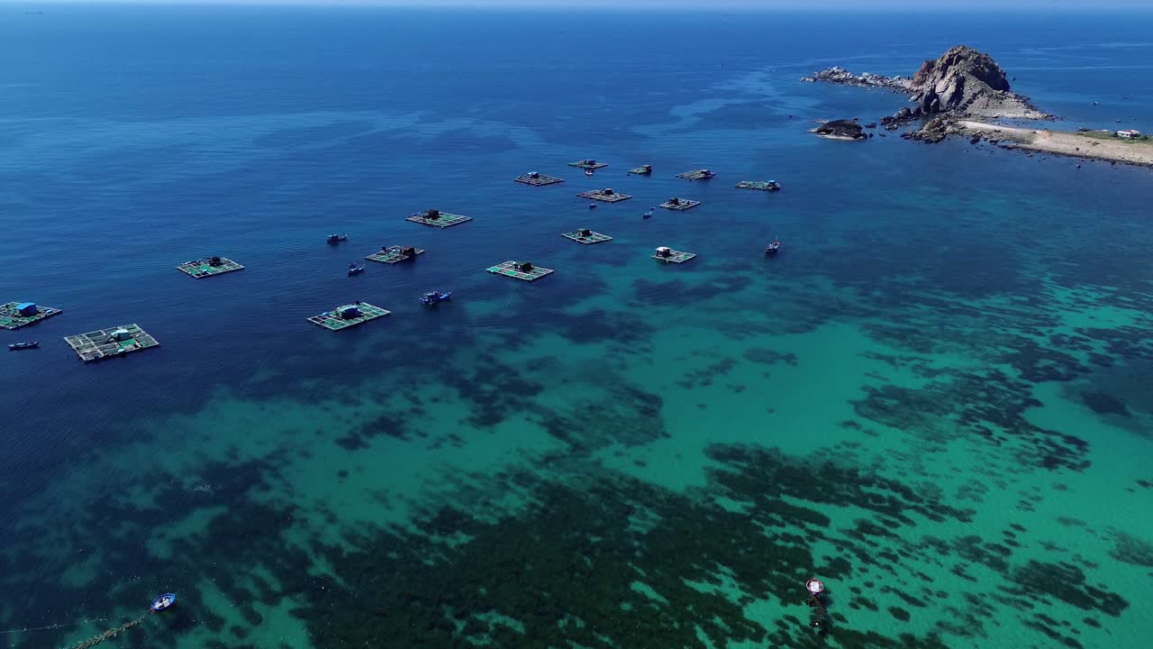 Aerial view of fishing village in Ninh Hải District, Ninh Thuận, moving slowly forward above fish farming platforms set in vibrant blue waters.