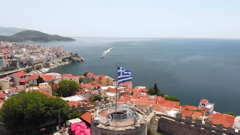 Aerial-Pull-Back-Shot-of-Kavala-Old-Town-Greece-and-Fortress,-Greek-Flag-Waving-on-Top-of-Tower-in-the-Castle