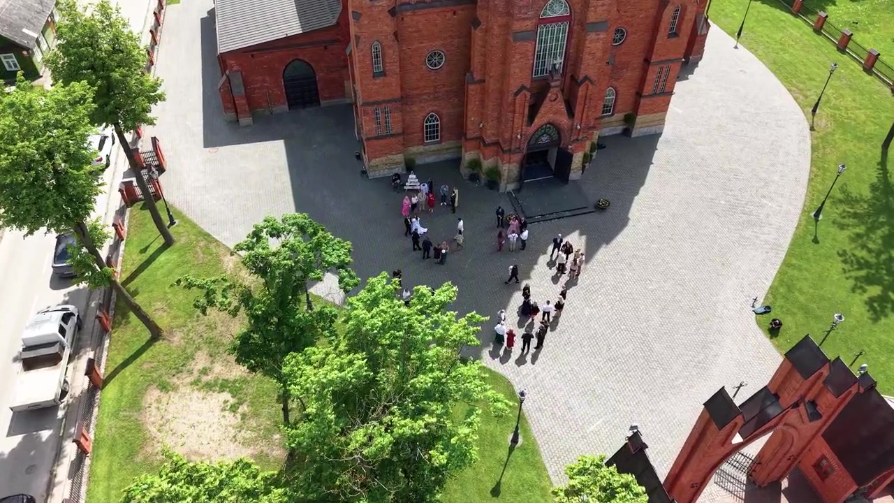 ドローン映像が上空からカイシャドリス大聖堂を映し出し、その赤レンガのゴシック建築、緑豊かな景観、外に集まる人々のグループ、そして明るい日差しが落とす鮮やかな影を際立たせる。