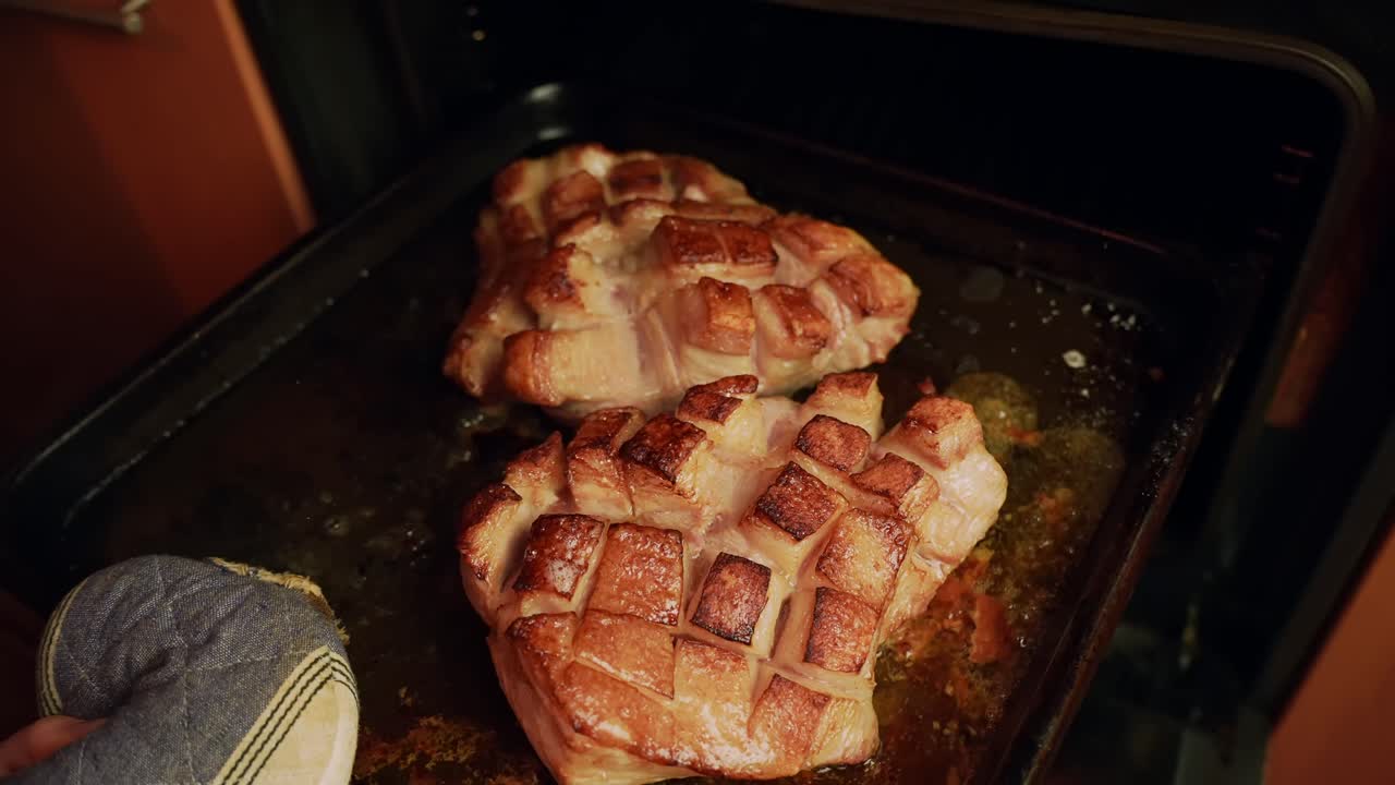 Un guanto da forno estrae una teglia calda dal forno, rivelando pancetta di maiale croccante con cotenna incisa e scoppiettante, e sughi sfrigolanti in una cucina domestica.
