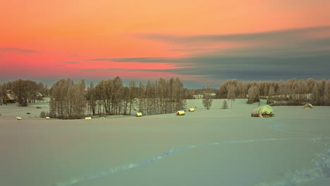 Aerial-time-lapse-shot-of-flying-clouds-on-colorful-sky-early-in-the-morning-in-winter---Snow-covered-hay-bales-on-countryside-fields