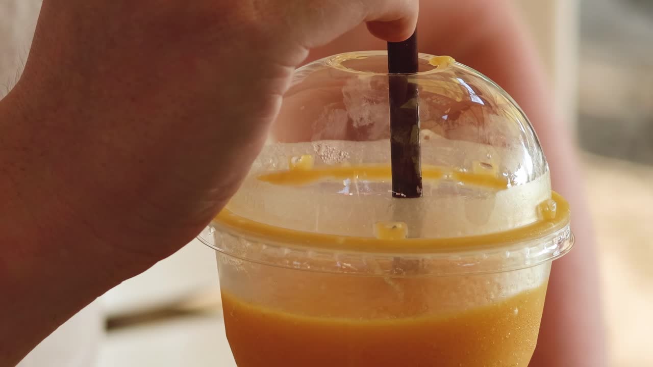 Una mano mescola una bevanda all'arancia con una cannuccia in un bicchiere di plastica con coperchio a cupola