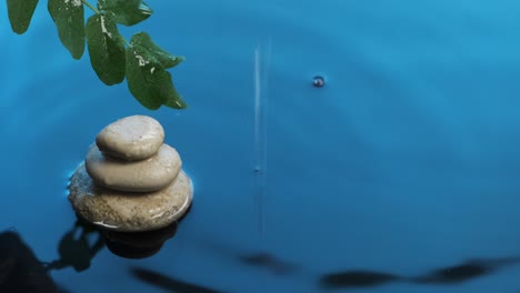 falling drops of rain into blue water. pyramid made of white stones and a branch of green leaves above the water.