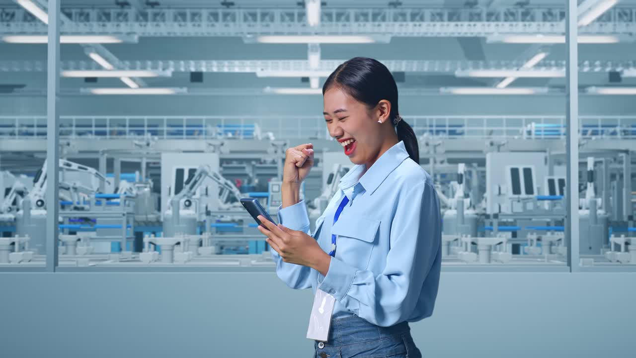 Side View Of An Asian Female Professional Worker Standing With Her Smartphone at Factory Digitalization for Innovative Project, She Raises Her Fist Up With Screaming Goal After Check On The Smartphone