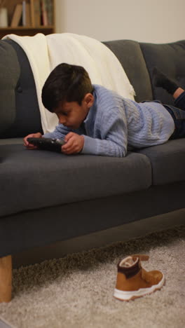 vertical video of young boy lying on sofa at home playing game or streaming onto handheld gaming device