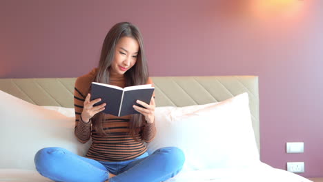 close-up of a young, attractive woman sitting cross-legged on a bed as she reads through a book