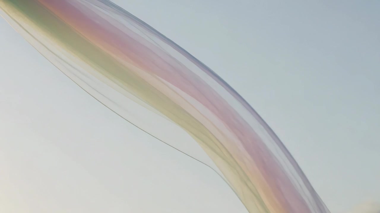 Drifting iridescent ribbon curling across blue sky, pushed by breeze with rainbow bands