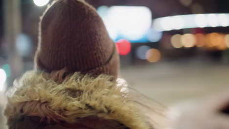 first-person view of a woman in a winter coat with a fur hood, standing still at night with blurry moving cars and colorful lights in the background