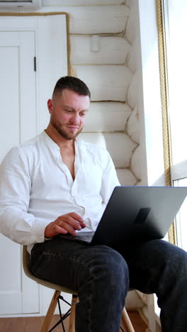 bearded man wearing white shirt and black jeans sits on the chair. freelancer works on his laptop sitting at the window. vertical video.
