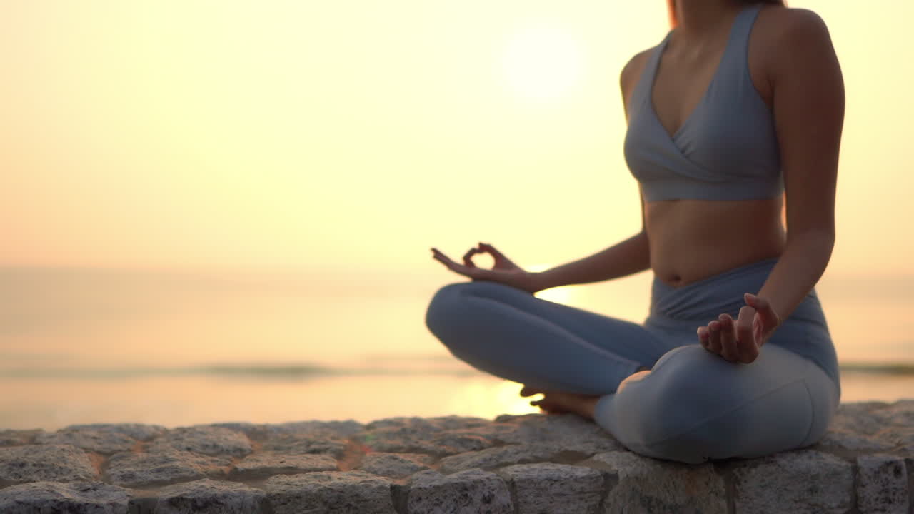 Static shot of a fit sexy lady doing yoga and meditation on a stone fence next to the ocean during sunset