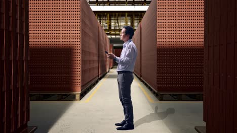 full body side view of asian male worker standing with his phone with red brick packed in stacks are stored, he observes by looking around before he come to concentrating with his smartphone
