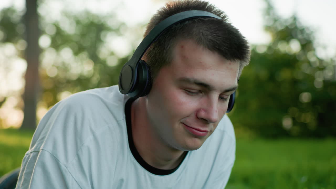 primer plano de un joven artista acostado en la hierba con auriculares asintiendo a la música mientras usa el teléfono y mirando hacia arriba con una sonrisa sutil, rodeado de vegetación y fondo borroso suave