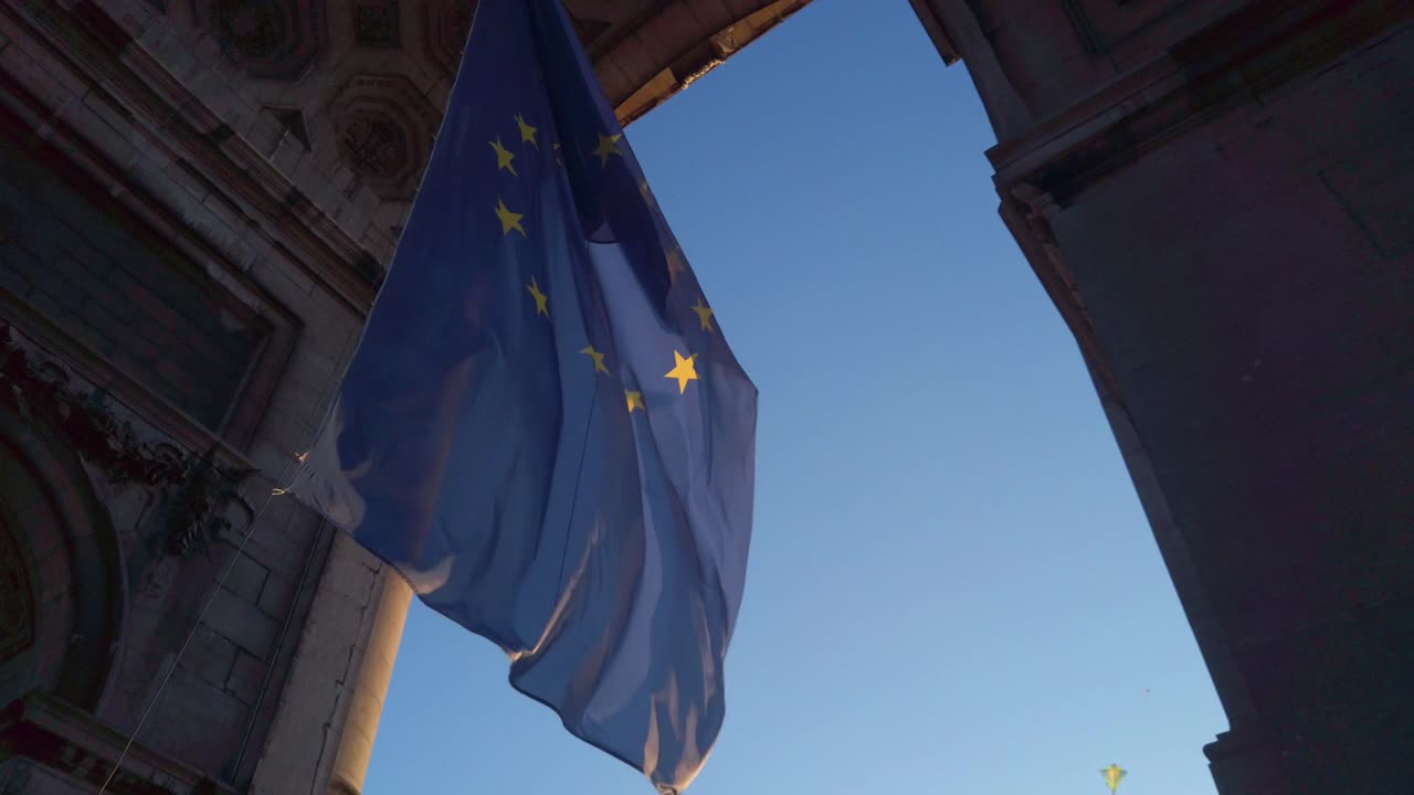 Emblema europeo che ondeggia lentamente sotto un grande arco di pietra contro un cielo pomeridiano di un blu intenso.