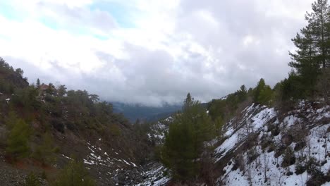 Drone-shot-entering-snowed-valley-through-trees-revealing-mountains-clouds-on-horizon