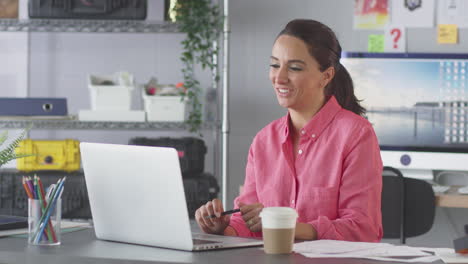 Businesswoman-In-Office-Making-Video-Call-At-Desk-On-Laptop-Showing-Designs-To-Client