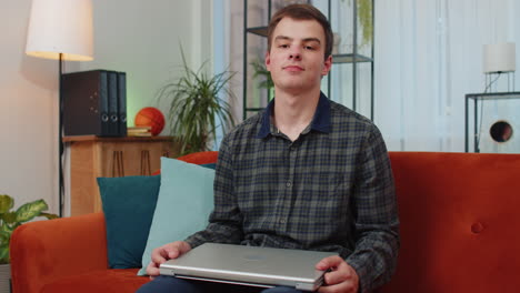 Young-man-freelancer-sitting-on-sofa-closing-laptop-pc-after-finishing-work-in-living-room-at-home