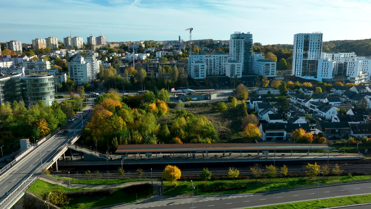 Aerial view of a city district with modern buildings, autumn trees and a wide road leading toward residential blocks under a clear blue sky