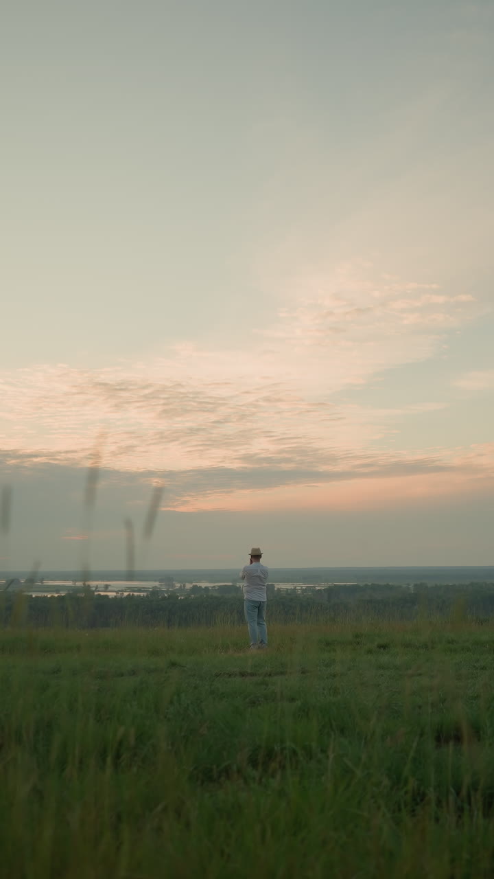 una vista lejana de un hombre vestido con una camisa blanca, sombrero y vaqueros, de pie contemplativamente en un campo cubierto de hierba junto a un lago tranquilo al atardecer.