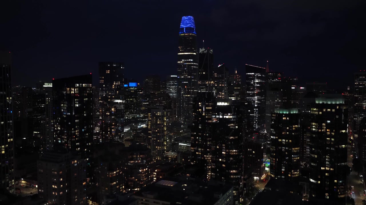 Downtown skyline illuminated at night with glowing skyscrapers, city lights and dark sky, iconic blue-lit tower rising above dense urban core. San Francisco, California. Aerial rising wide shot. Quiet