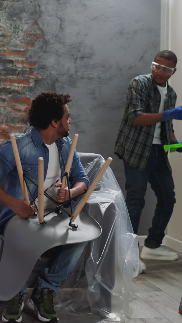 Happy African-American guy paints wall and asks for point of view of brother fixing chair in living room. Black workers do repair together in apartment