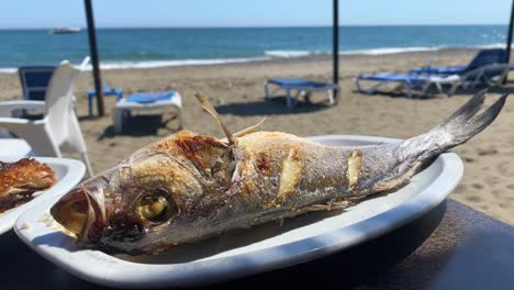 Pescado-De-Lubina-A-La-Parrilla-En-Un-Plato-En-La-Playa,-Comida-Tradicional-Española-En-Un-Restaurante-Con-Una-Hermosa-Vista-Al-Mar,-Día-Soleado-En-Marbella-España,-Toma-De-4k