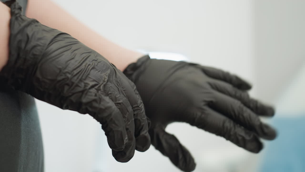 Close-up of beautician arranging black glove on hand while preparing for work, dressed in dark apron. Scene shows careful hand movement with soft background blur in clean, well-lit salon setting