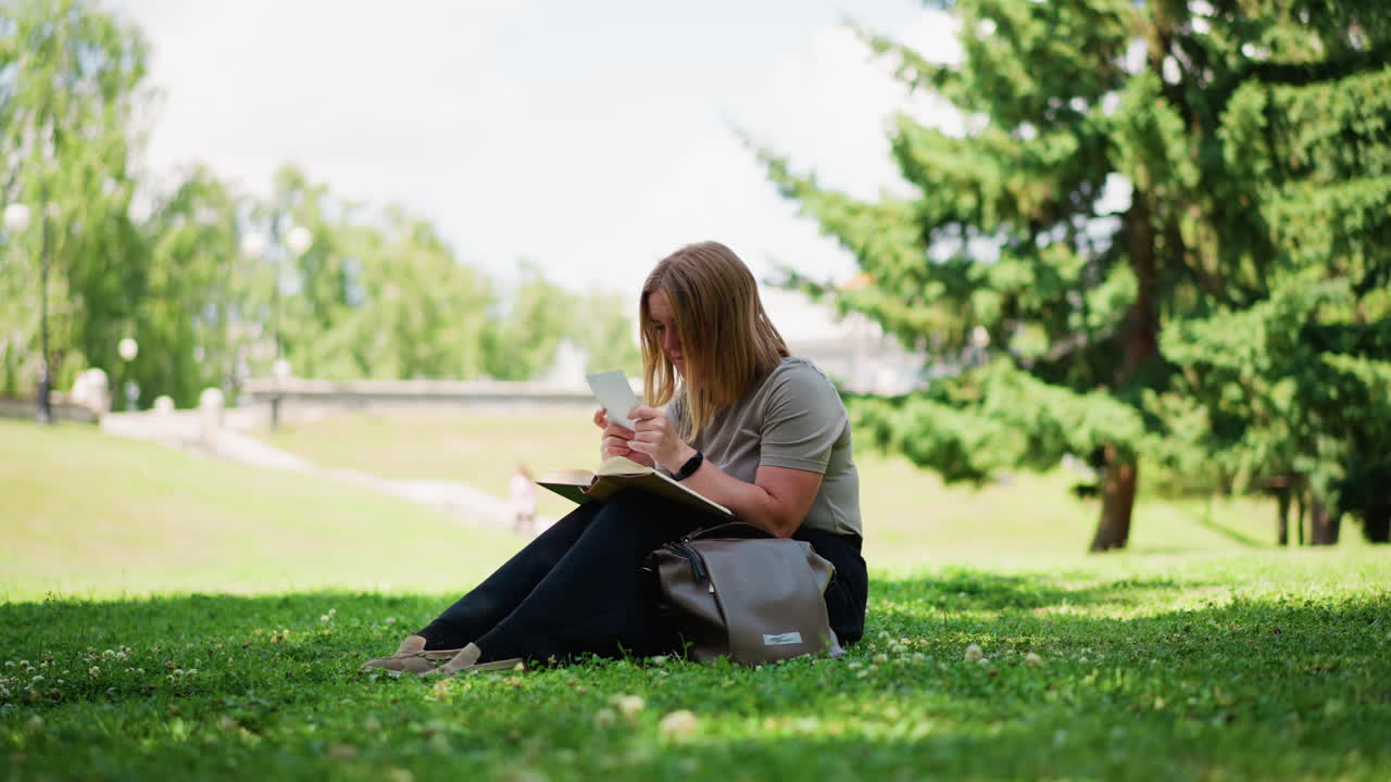 Young girl sitting on green grass under trees flipping through old book discovering hidden photo, looking at it with sadness and reflection, calm summer atmosphere filled with emotion