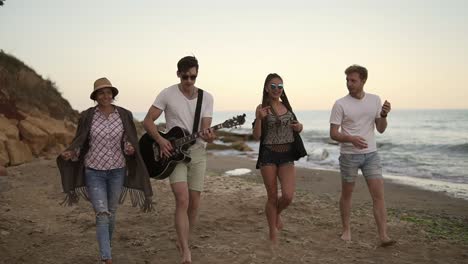 young cheerful happy friends walking on the sand by the ocean, playing guitar, singing songs and dancing. slow motion shot