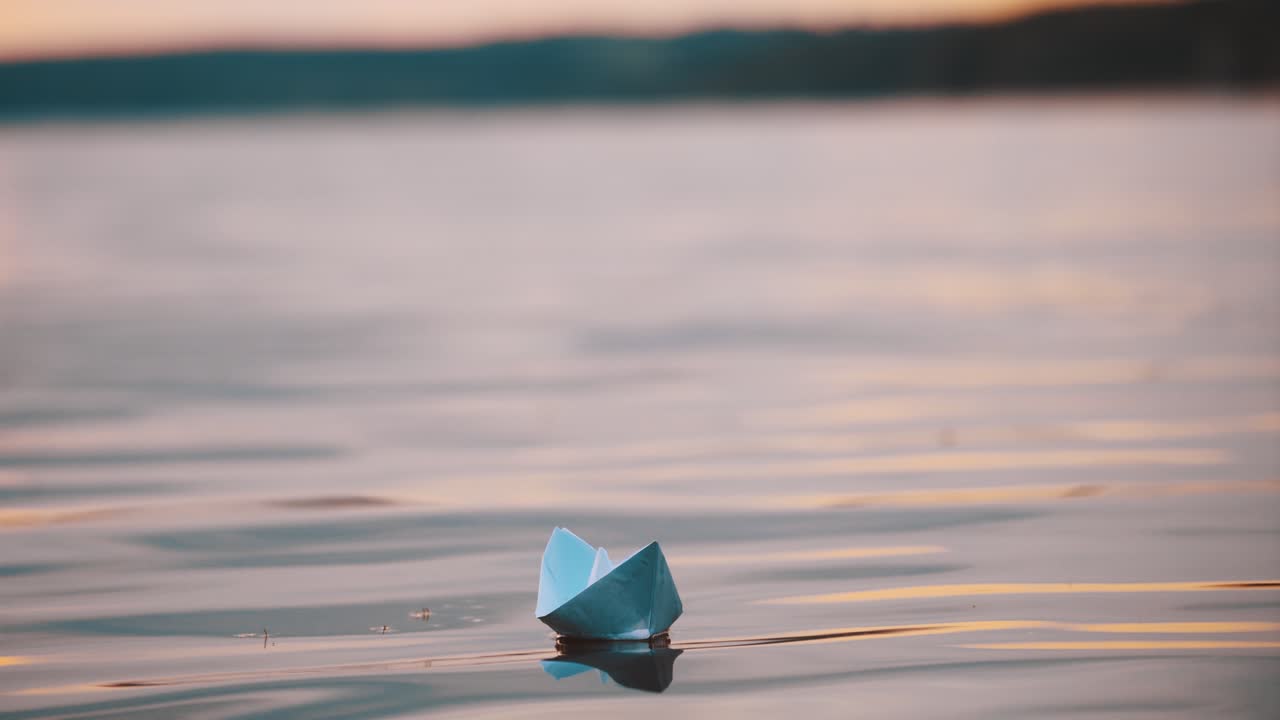 Blue homemade boat on water on blurred evening background. Paper little ship floating alone in calm river at sunset.