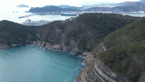 drone aerial view in south korea countryside flying over the clear blue sea of busan next to a green mountain, ships and city in the background sunny day