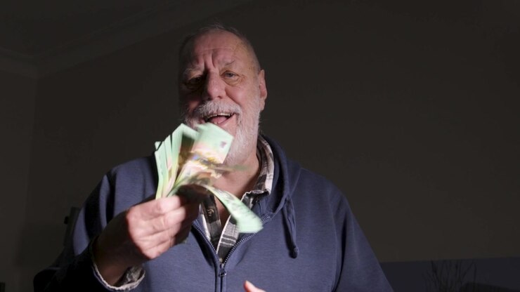 Un uomo anziano con felpa con cappuccio sventola una spessa pila di banconote verdi sotto una dura illuminazione laterale, suggerendo stress finanziario personale o una vincita inaspettata