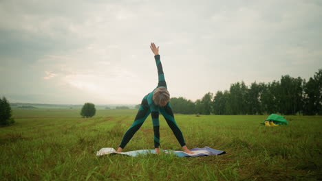 woman in green and black suit on yoga mat practicing triangle forward pose in serene grassy field under cloudy skies with trees in the background, arms stretched wide