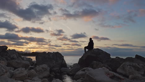 Timelapse-captures-man-sitting-on-rocks-experiencing-beautiful-golden-sunrise-by-the-seaside-in-Sitges,-Spain