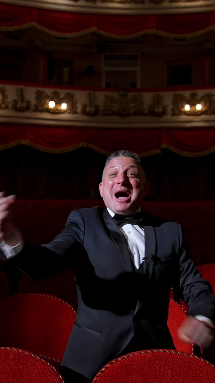 Happy actor in dark theatrical hall. Talented man in suit standing among vacant red chairs and shouting cheerfully. Portrait of a comedian in empty theater. Vertical video