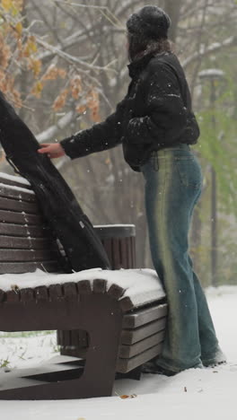 dama quitando el paquete de guitarra para colocar en el banco cubierto de nieve en un parque sereno, mientras que otra persona camina en el fondo nevado lejano