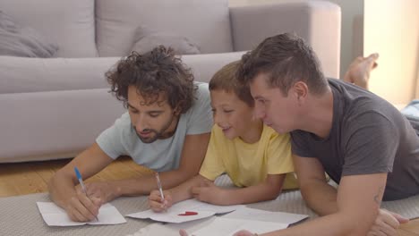 cheerful dads and son lying on floor and drawing in papers