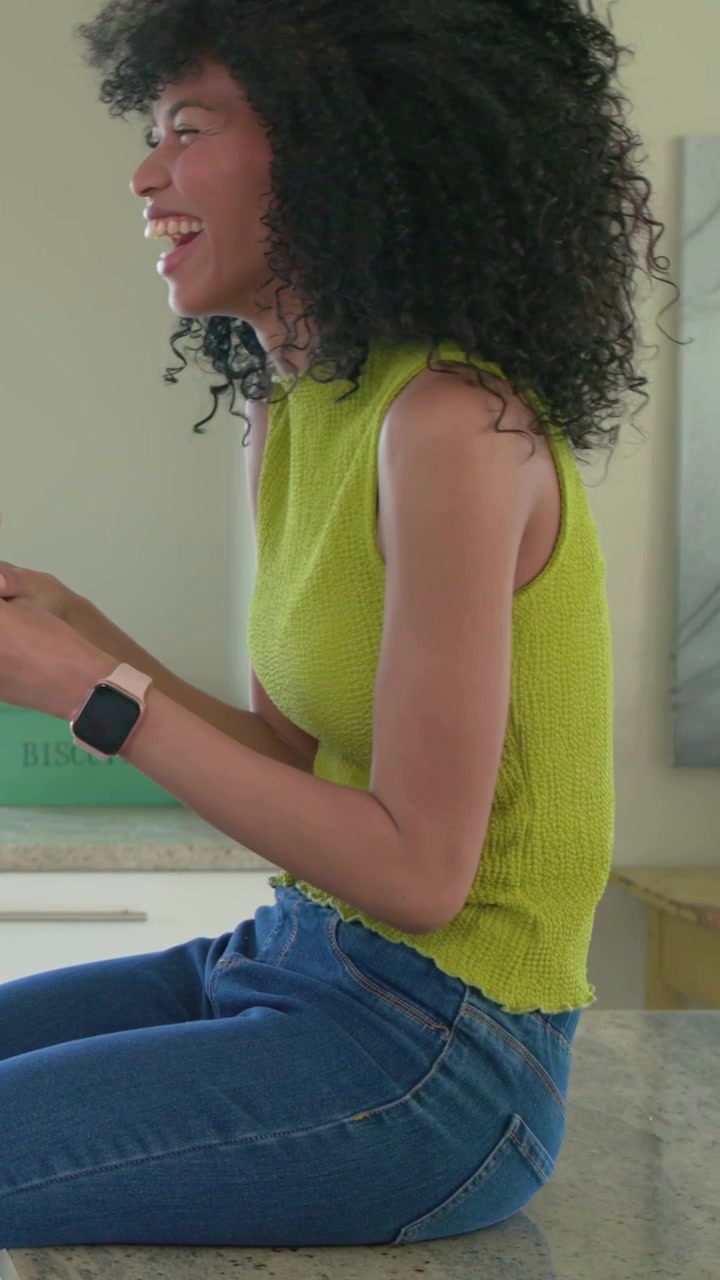 Video verticale: Donna con top color lime che si allunga per prendere una tazza da sinistra sul bancone della cucina, sorseggiando per chiacchierare