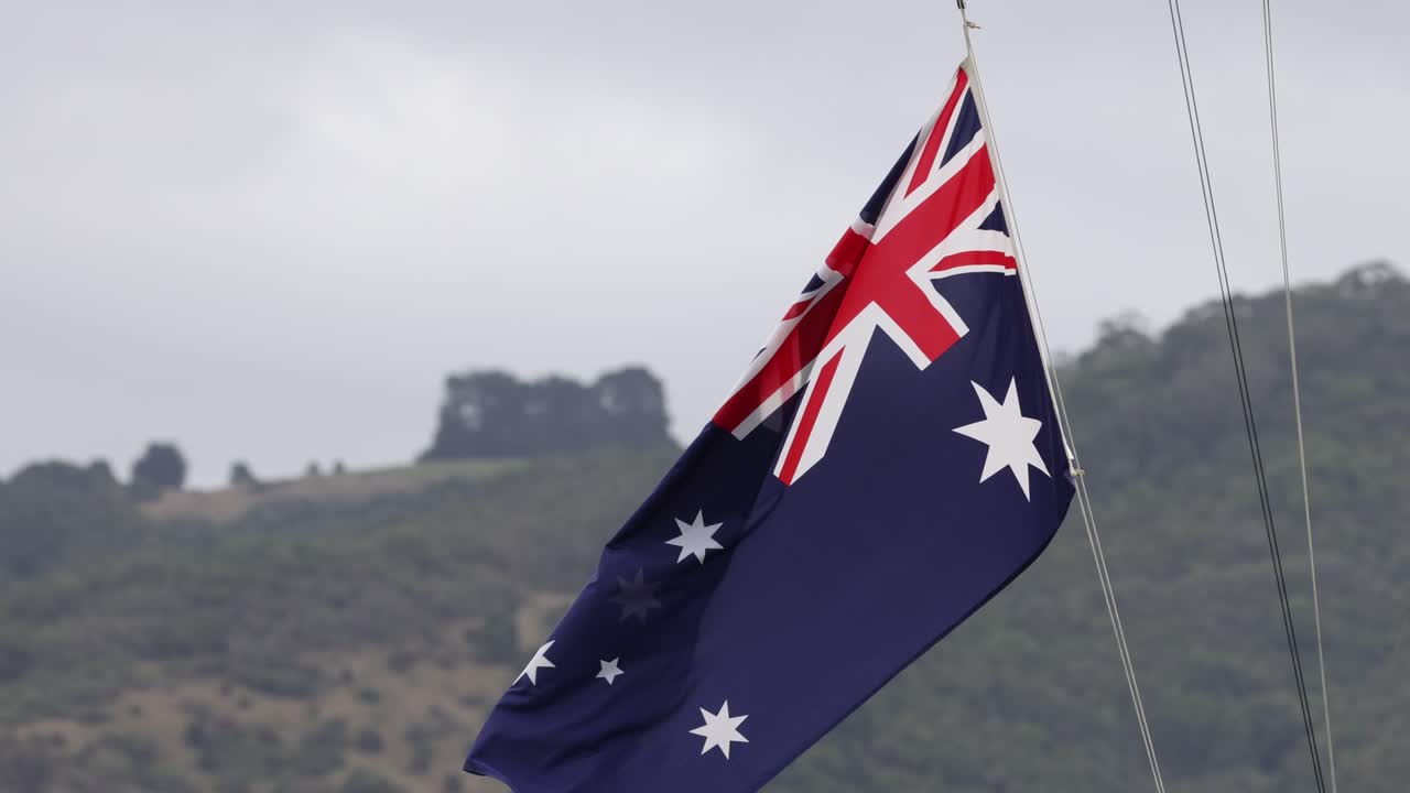The Australian flag waves in the wind against a hilly backdrop on a cloudy day at Great Ocean Road