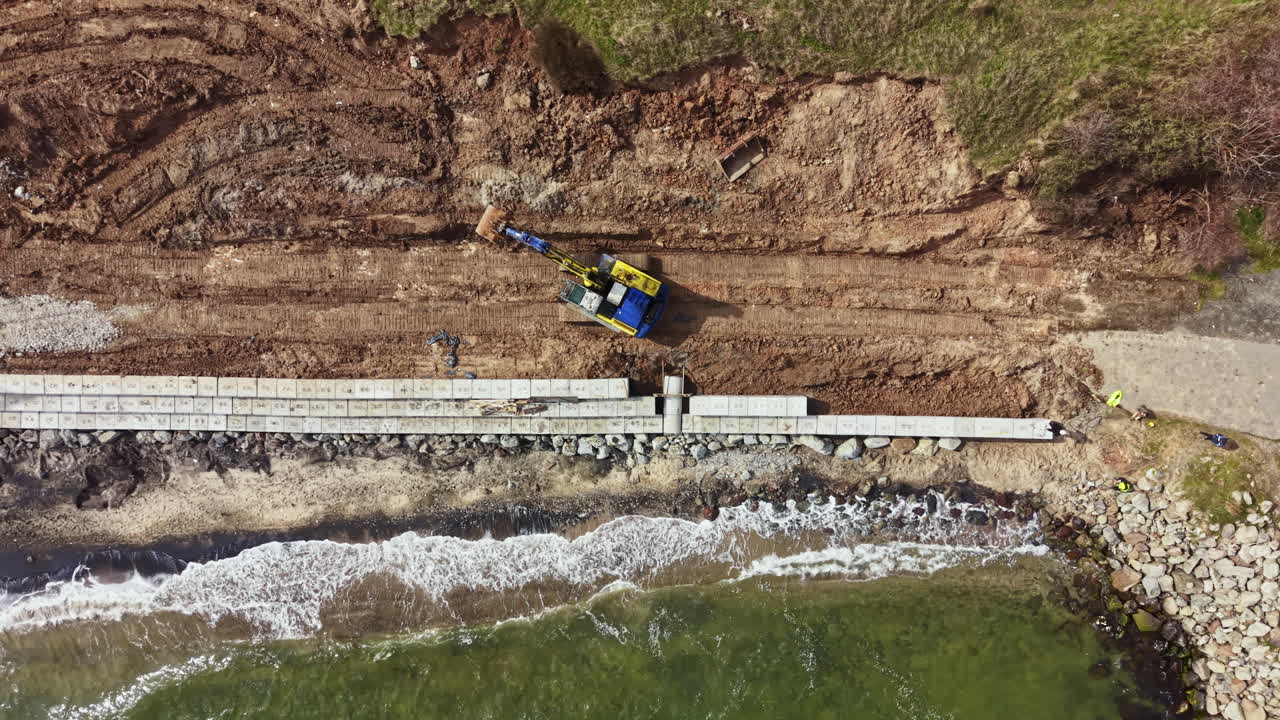Lavori di costruzione in un'area costiera con attrezzature e operai coinvolti nella realizzazione di una barriera vicino alla riva durante il giorno.