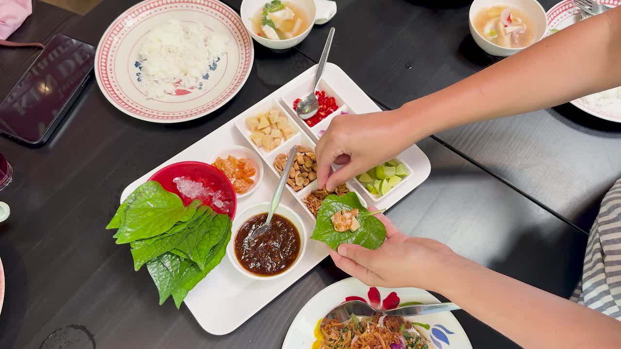 Hands prepare traditional Miang Kham by adding assorted fillings to betel leaves on a white tray, under bright, even lighting, viewed from overhead